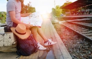 Foto de uma estação de trem. Em primeiro plano, uma pessoa sentada com uma mochila, um chapéu, um mapa e uma câmera. Em segundo plano, outras pessoas na estação, a linha de ferro, árvores e o céu.