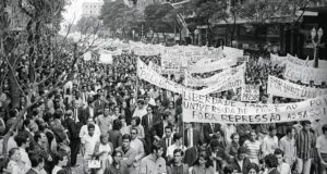 Foto em preto e branco mostrando a passeata dos cem mil em 1968. Na imagem, as ruas estão tomadas por pessoas com faixas pedindo liberdade