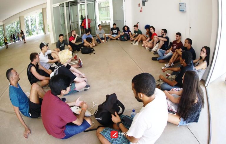 Foto de um grupo de cerca de 20 jovens sentados em um saguão formando um círculo para a discussão de obras vistas no Instituto Inhotim.