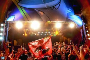 Foto de um palco com uma e uma plateia na parte inferior da imagem. Ao centro, vê-se uma bandeira vermelha com o escrito “Show Medicina”, com o símbolo do Conclave Médico Desportivo da UFMG e com o mascote deles chamado Conclito.
