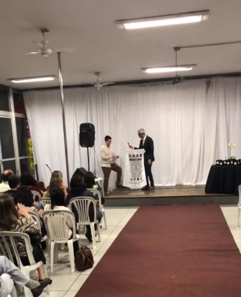 Foto da Homenagem dos Professores, no palco um aluno entrega o microfone para um professor falar no pupito. Evento realizado na sede do DAAB