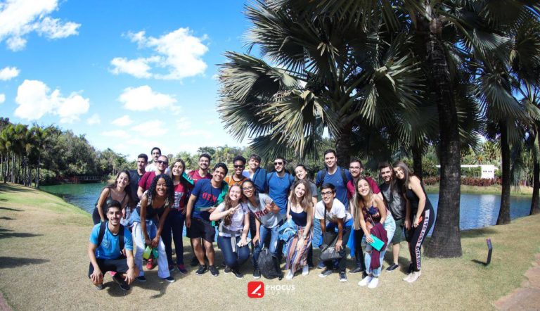 Foto de um grupo de aproximadamente 24 jovens juntos, em pé, de costas para uma lagoa, com algumas palmeiras no canto direito no Instituto Inhotim.