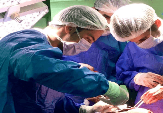 Foto com destaque para um estudante com capote e touca auxiliando numa operação