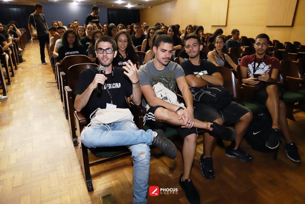 Foto de vários alunos sentados em uma plateia. Em evidência na imagem, um aluno com um microfone em mãos realizando uma fala.