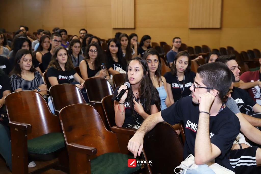Foto de vários alunos sentados em uma plateia. Em evidência na imagem, uma aluna com um microfone em mãos realizando uma fala.