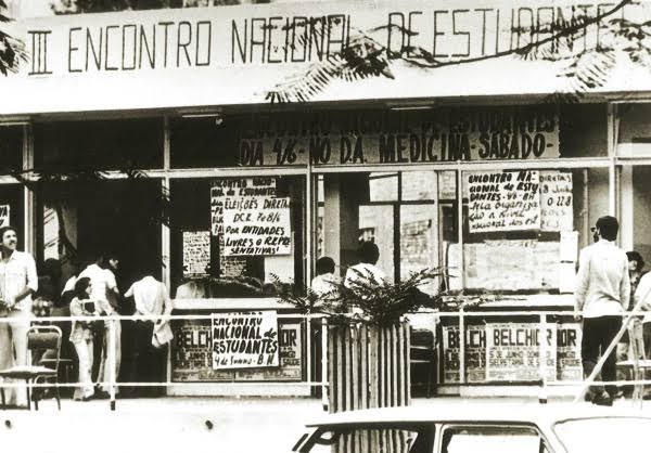 Foto em preto e branco do DAAB de 1977, na ocasião acontecia o 3º Encontro Nacional dos Estudantes. Na imagem, a fachada do DAAB está cheia de cartazes com palavras de ordem contra a opressão dos militares. Na porta do DAAB tem alguns estudantes.