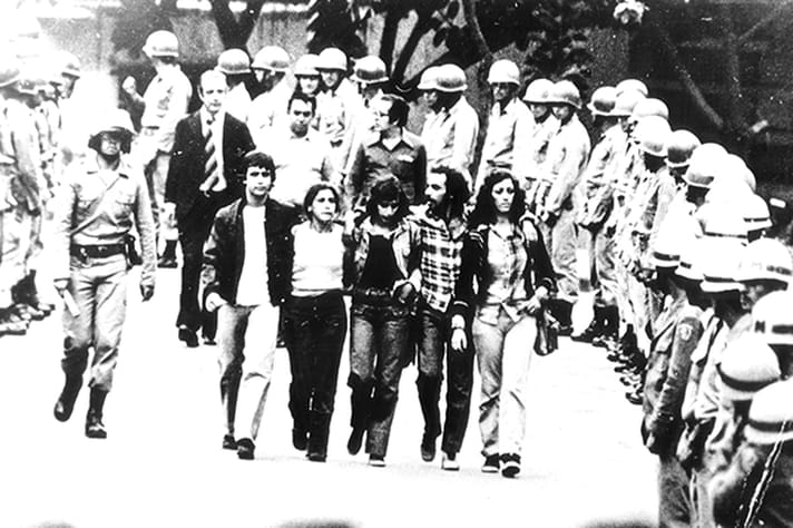 Foto em preto e branco tirada em 1977 no Campus Saude da UFMG. Na foto, há 5 estudantes caminhando juntos abraçados do DAAB para fora do Campus. Acompanhando o trajeto dos estudantes, há dezenas de militares guiando os estudantes para fora