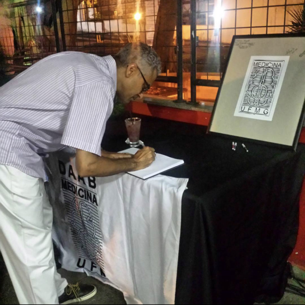 Professor Humberto em evento realizado no DAAB, 2018. Ele está assinando uma folha em mesa coberta com bandeira do DAAB.